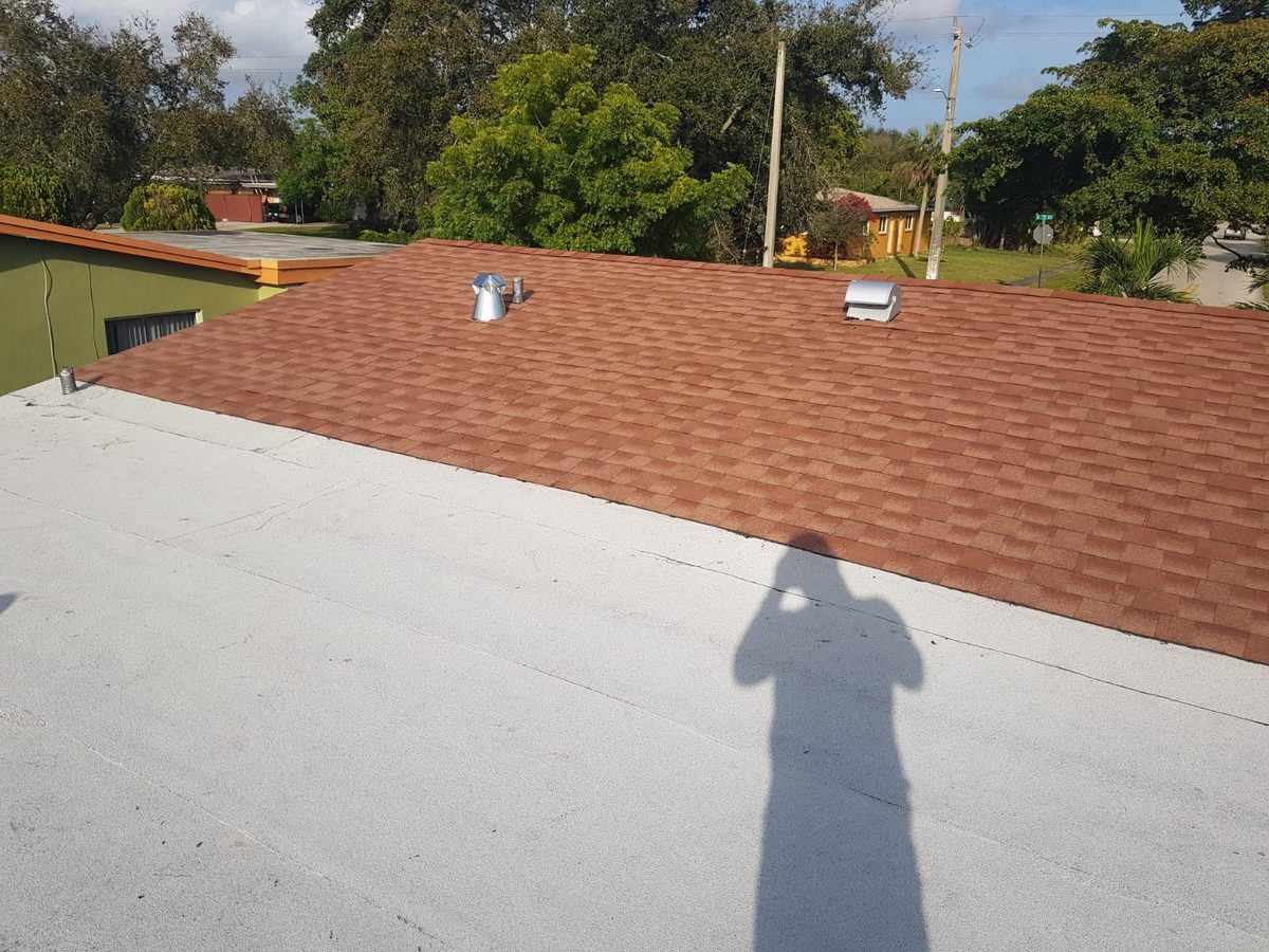 An overhead view of a residential roof with a flat white section in the foreground and brown shingles in the background.