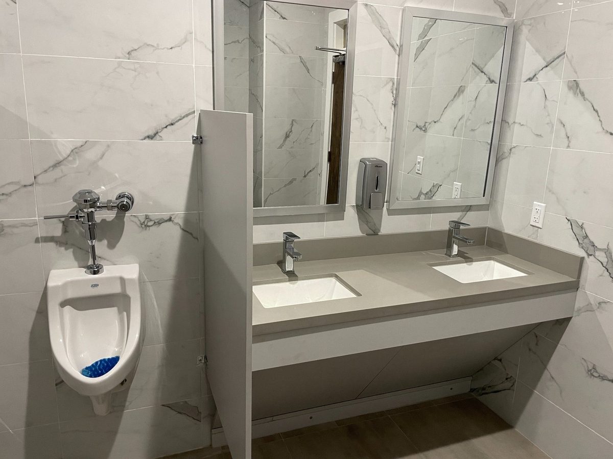 A commercial bathroom with a double vanity, mirrors, and a urinal against marble-look tiled walls.