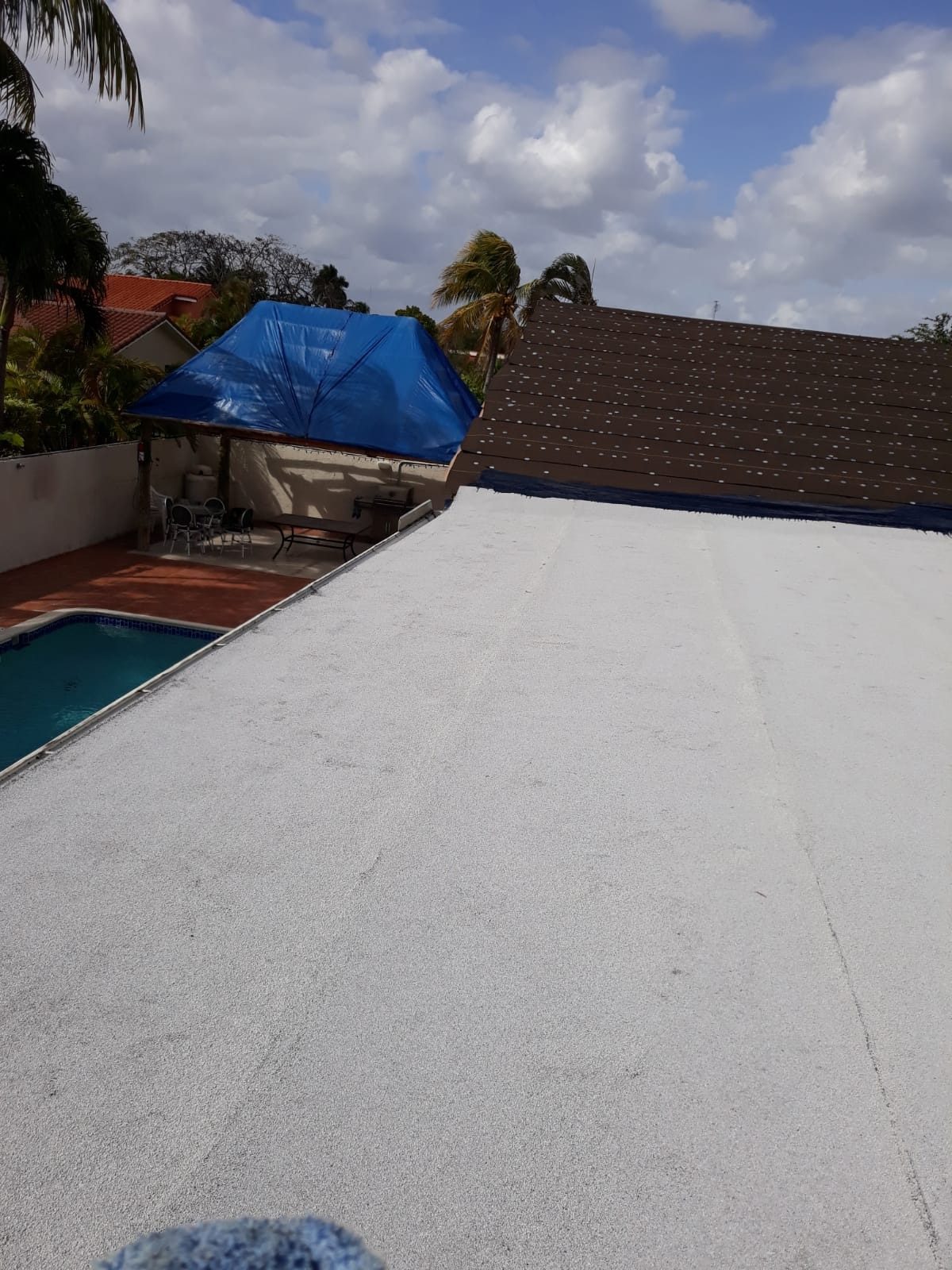 View of a finished white flat roof next to a brown shingle roof with a pool in the background.